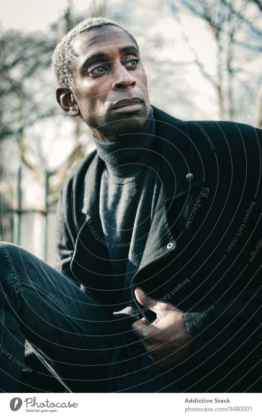 Trendy African American man in park style rest city adjust jacket ethnic urban modern male lifestyle casual trendy serious confident sit cool street guy