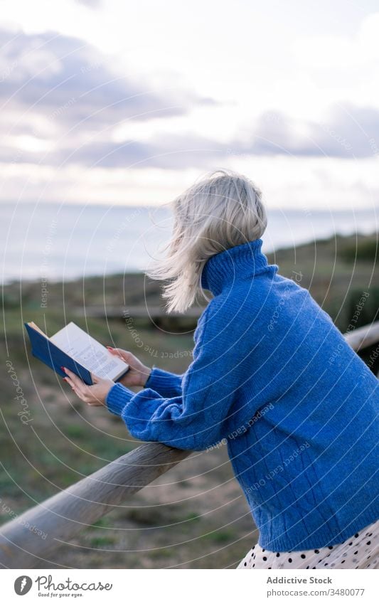 Young woman reading book near sea fence beach casual rest young blue enjoy poetry sweater nature relax female wooden style alone modern trendy blond recreation