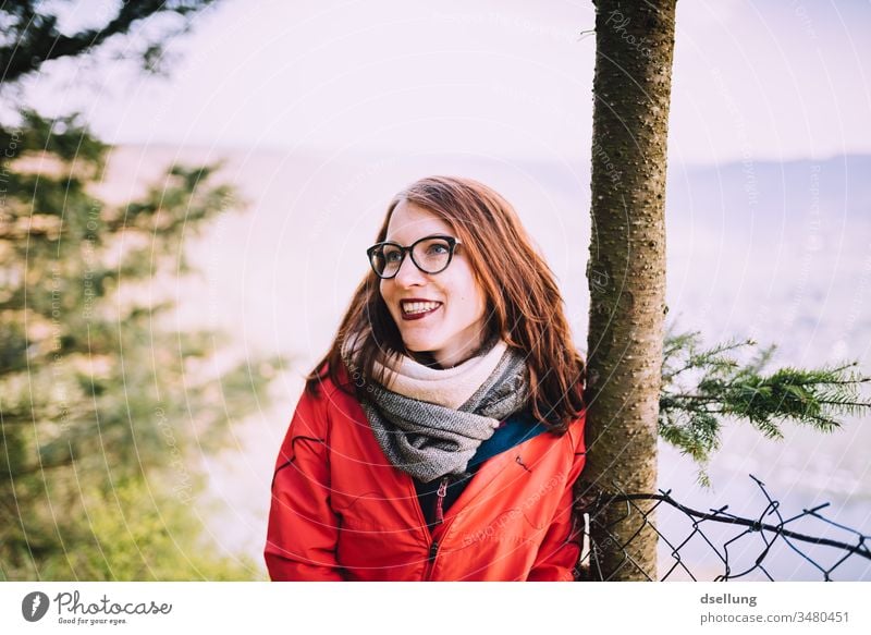 Young woman with a red jacket leans relaxed against a tree and smiles with the sun take a walk To go for a walk Adventure Healthy gap Hill Hiking Tourism