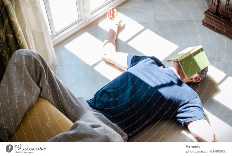 Young man resting on the floor whit a book on his face while enjoying the sun coming through the window. Concept of stay at home, freedom, boredom... leisure