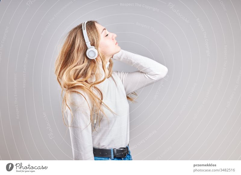 Young woman girl listening to music streaming content having fun watching video enjoying video chat talking with friends making gestures faces using smartphone earphones headphones standing over plain grey background