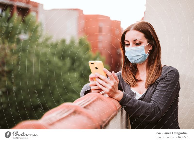 young woman at home on a terrace wearing protective mask, using mobile phone and enjoying a sunny day. Corona virus Covid-19 concept smart phone technology