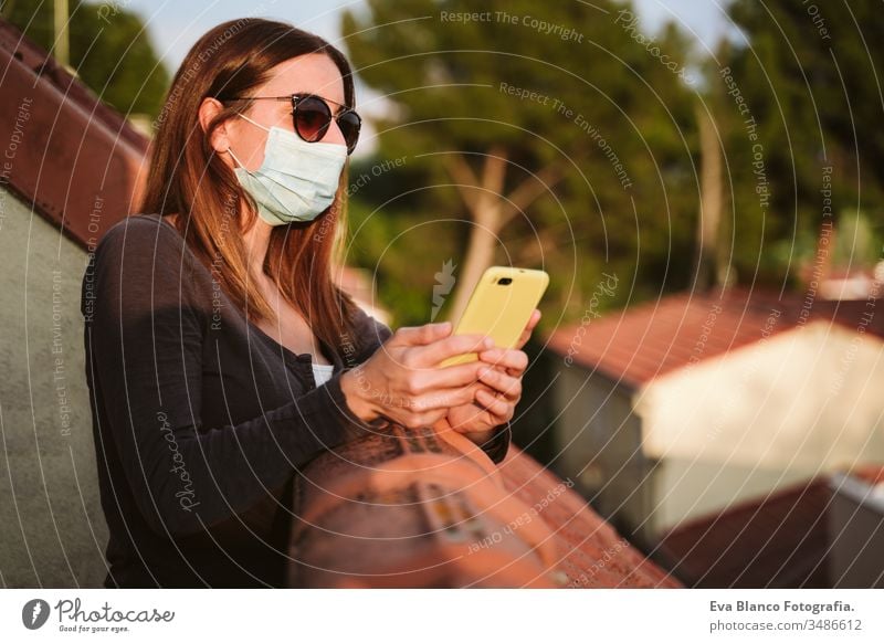 young woman at home on a terrace wearing protective mask, using mobile phone and enjoying a sunny day. Corona virus Covid-19 concept smart phone technology