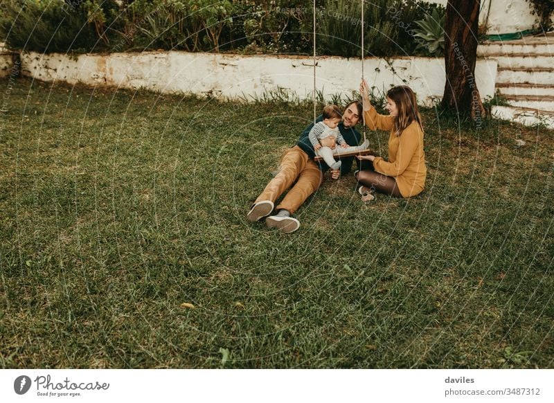 Beautiful couple enjoying with their baby son in a swing, on the green grass of the garden at the countryside excitement enjoyment smiling sitting care innocent