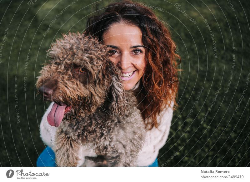 A beautiful woman is in the meadow with her dog. The owner is hugging her pet while she is looking at the camera. They are enjoying a day in the park. The pet is a Spanish water dog with brown fur.