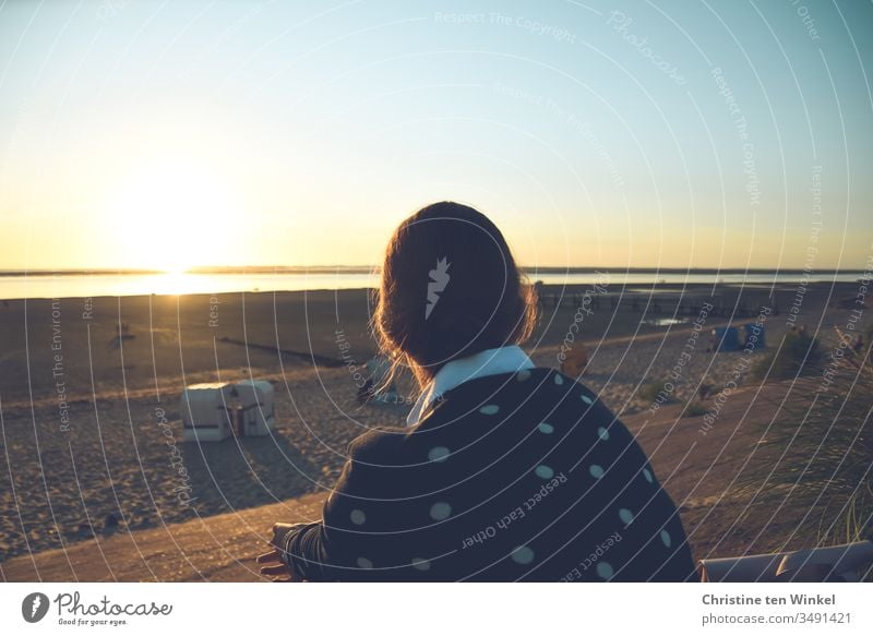 Young woman sits with her back to the camera on the beach promenade and looks over the beach into the sunset over the North Sea Beach Sit Ocean