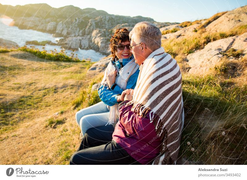 Loving mature couple hiking, sitting on windy top of rock, exploring. Active Mature man and woman wrapped in blanket, hugging and Happily smiling. Scenic view of sea, mountains. Norway, Lindesnes.