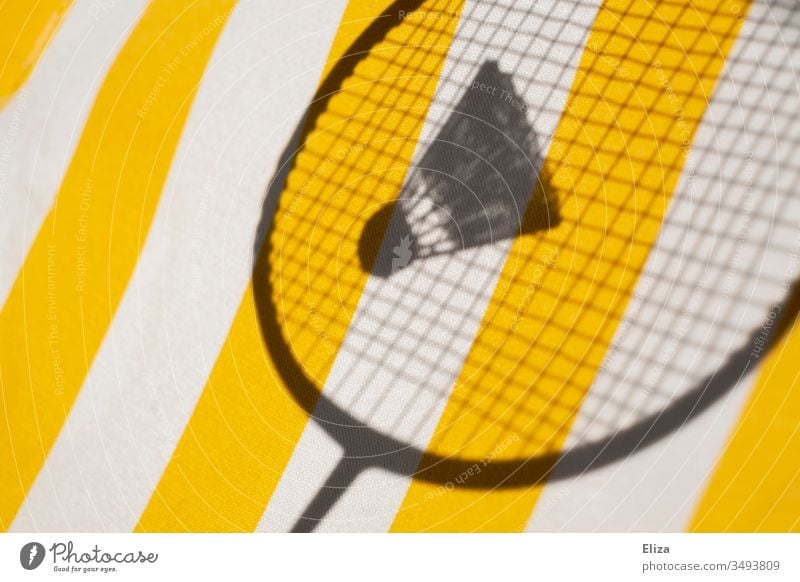 The shadow of a badminton racket with a shuttlecock in front of a striped yellow background; badminton Shuttlecock Badminton badminton rackets Badminton racket