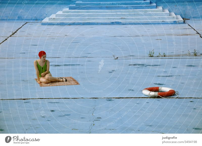 The girl with the beautiful red bathing cap and green swimsuit is sitting in an empty pool and looking at a life ring. A summer love. Girl Woman Swimwear