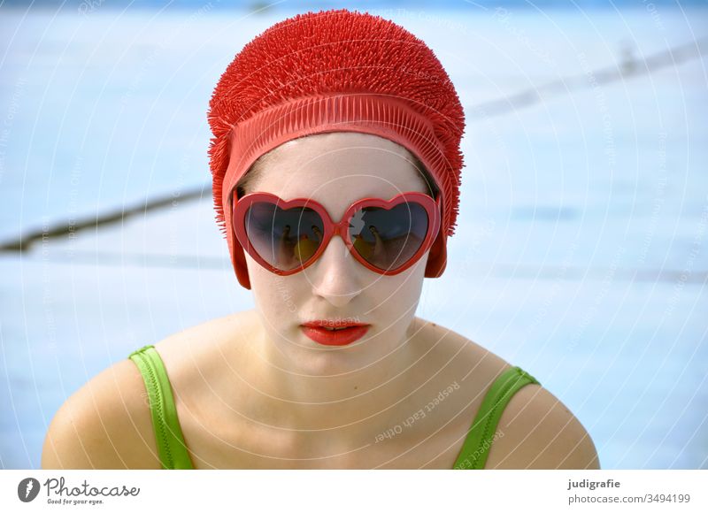 The girl with the beautiful red bathing cap and the green swimsuit looks through heart-shaped sunglasses directly into the camera. A summer love. Girl Woman