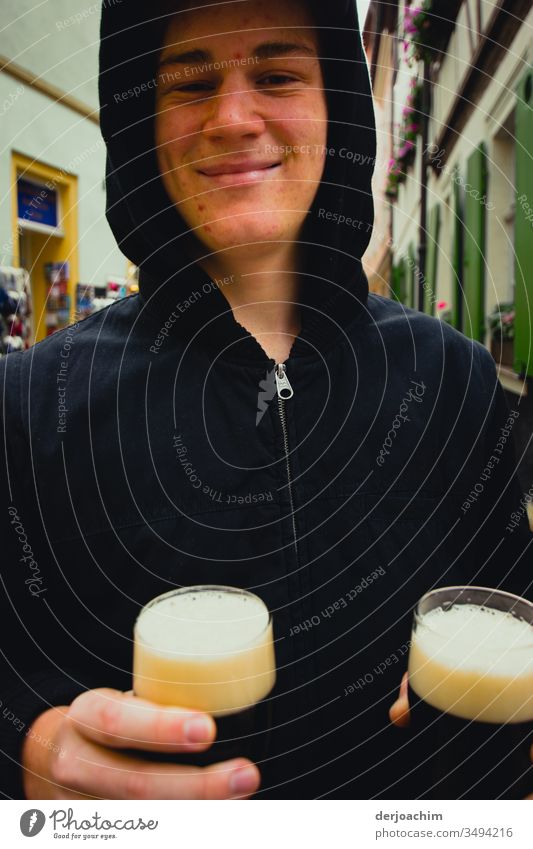 Young man holds two glasses with dark beer on the street ( smoked beer ) in his hand and smiles. The white foam in the glasses is well recognizable on his dark jacket.