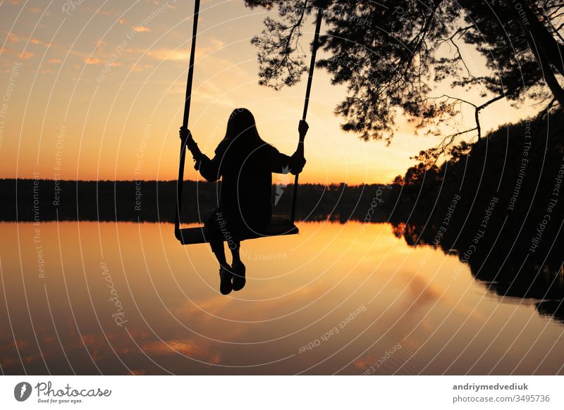 romantic young woman on a swing over lake at sunset. Young girl traveler sitting on the swing in beautiful nature, view on the lake swinging summer boy