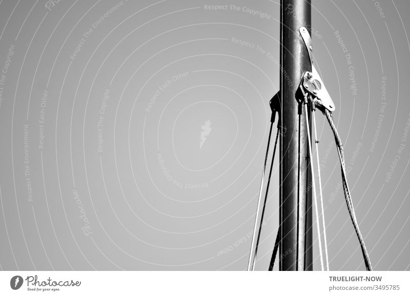The aluminium mast of an old Ixylon sailing dinghy with wall hangers and shrouds in partial view black and white against the neutral background of a cloudless sky
