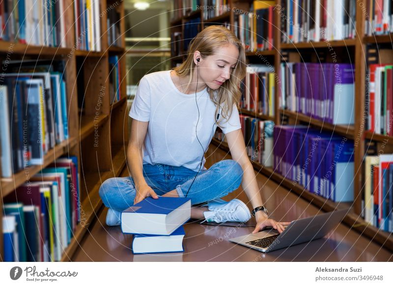 Young girl sitting on the floor in traditional library at bookshelves, reading books. Smiling and laughing student working with laptop, studying. Higher education.  Student life