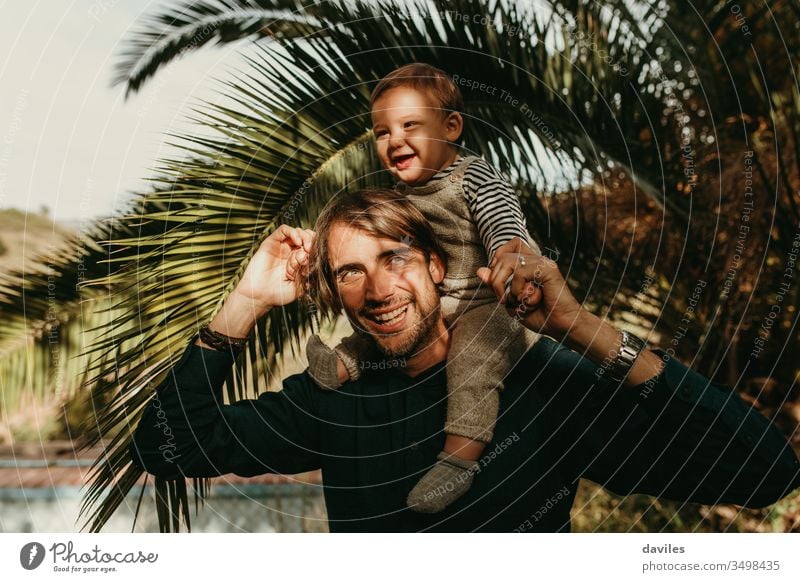 Laughing father giving his son a piggyback ride outdoors with sun light in the face. lovely smile white playing carrying shoulders handsome joyful hipster care