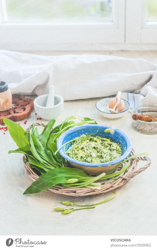Homemade tasty wild garlic butter in bowl with fresh ingredients on light kitchen table at window background. Seasonal cooking and eating homemade seasonal