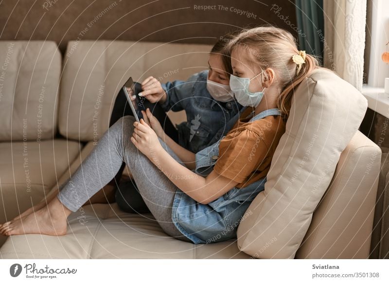 Two cute girls in medical masks are sitting on the sofa with a tablet. Schoolgirls do their homework. Distance learning during the COVID-19 quarantine.