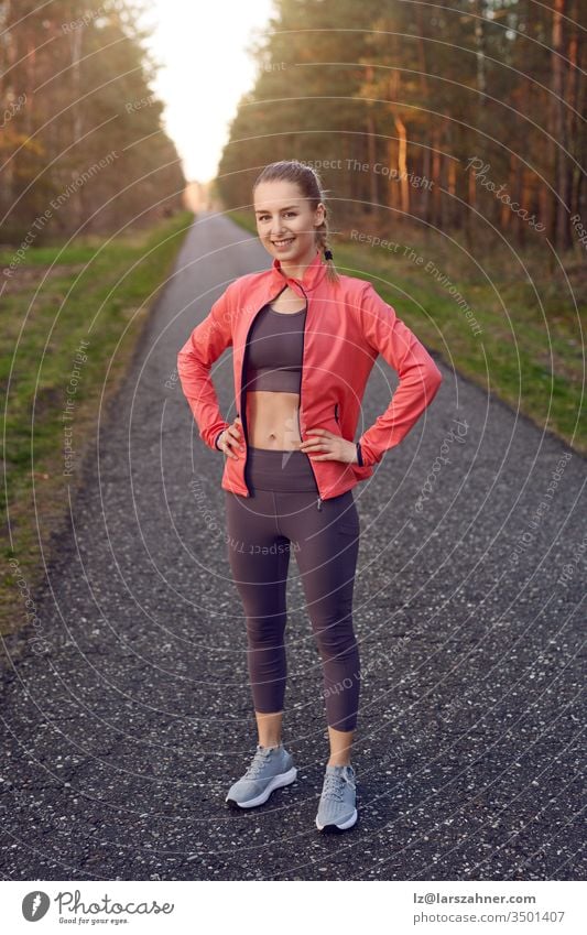 Full length front portrait of sporty young girl in training wear with grey pants and red jacket, standing and resting after jogging with her hands on hips, looking at camera and smiling