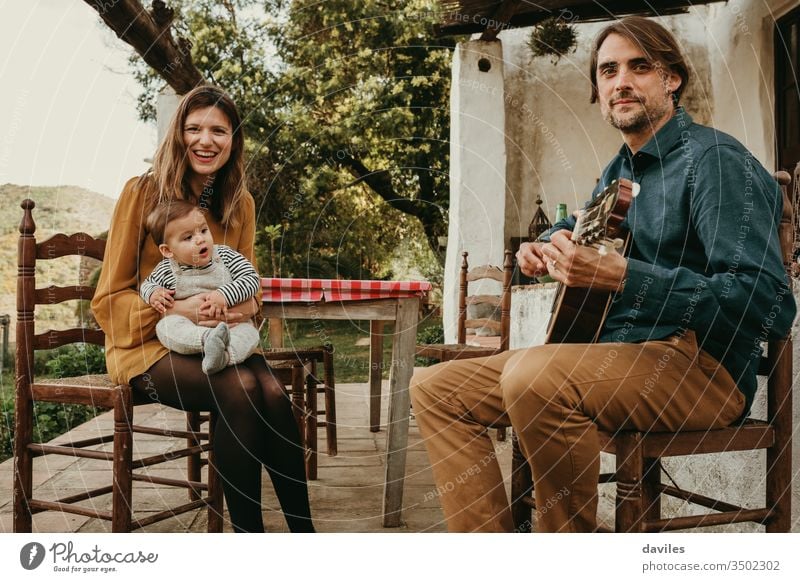 Beautiful couple with their baby infant enjoying sitting on the courtyard at countryside. Man is playing the guitar. Woman holds the baby. They look at the camera.