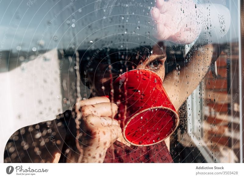 Thoughtful woman drinking coffee near window sad depression self isolation home unhappy lonely covid19 melancholy young female wet rain solitude stress cup