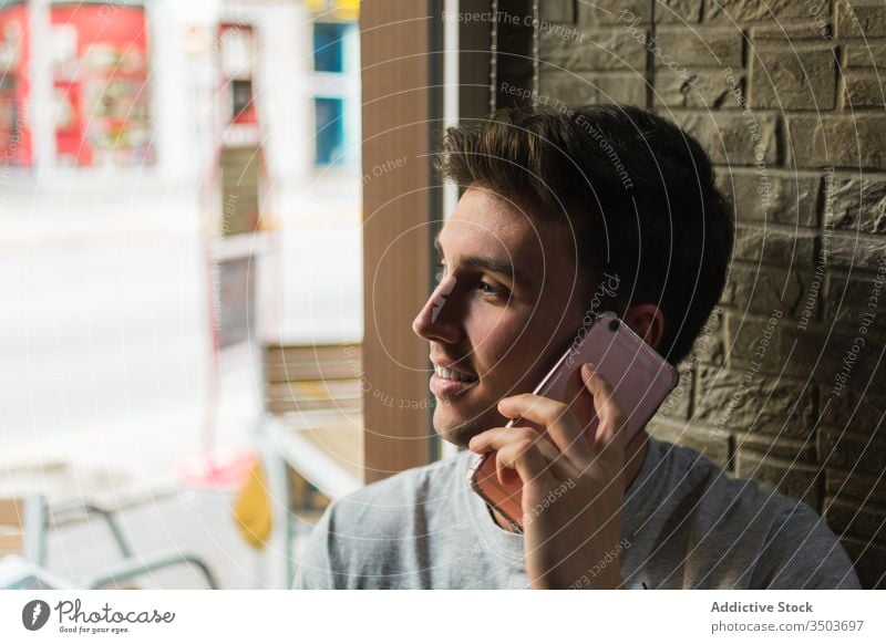Cheerful young man speaking on smartphone near cafe window smile modern happy call male lean restaurant mobile cheerful chat lifestyle relax guy conversation