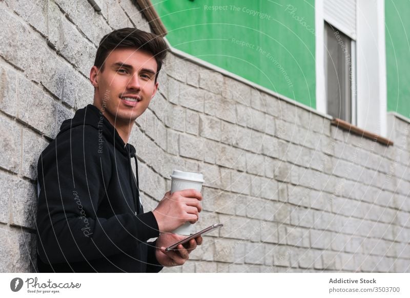 Cheerful man with smartphone and drink to go resting on street using city lean wall positive male building young takeaway beverage cup coffee tea cheerful