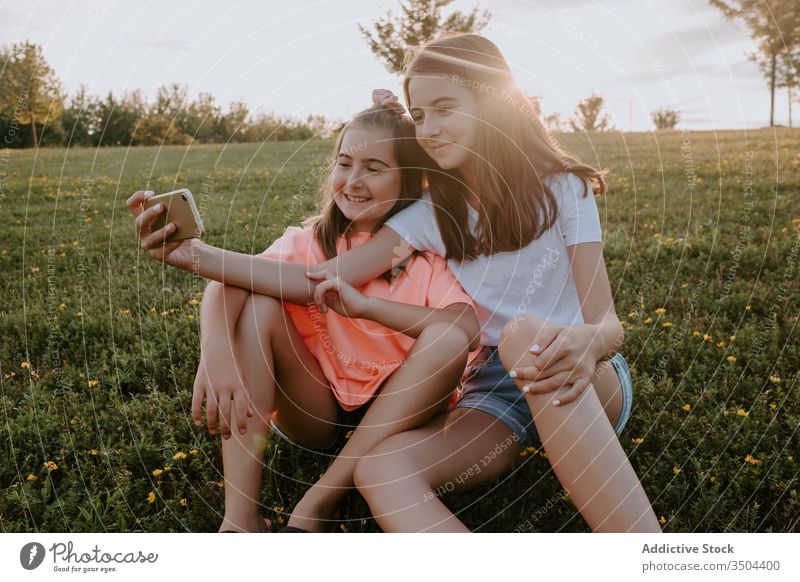 Adorable sisters sitting in a park and taking a selfie kid nature together happy hug grass summer girl countryside cheerful teen teenage green meadow sibling