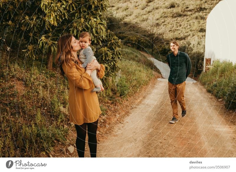 Woman kissing her baby son in nature, standing in a path, while man approaches to them with a smile. countryside loving family walk in nature tree happy parents