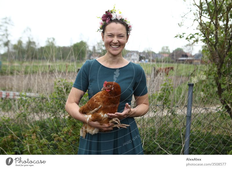 Young woman with wreath of flowers in her hair is standing in the garden holding a brown chicken in her arms Central perspective Shallow depth of field Day
