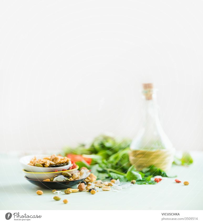 Healthy salad making ingredients on white table at light sunny wall background. Fresh kitchen herbs. Nuts topping. Olives oil. Salad dressing . Diet or vegetarian food concept.