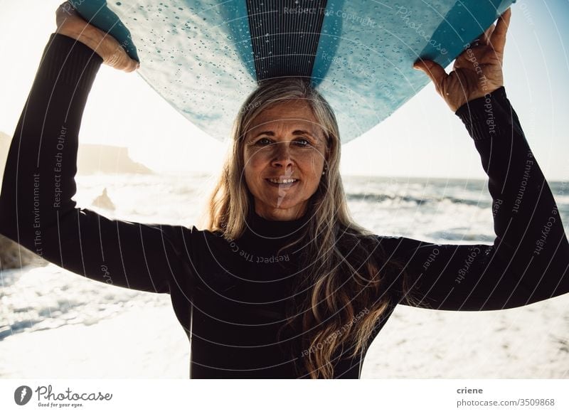 Smiling senior woman carrying surfboard at beach looking at camera women vacation surfing adult smile grey hair lifestyle joy caucasian sport hobby portrait
