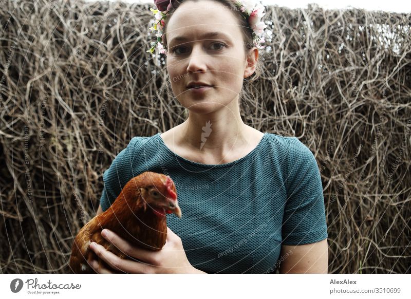Young woman with a wreath of flowers in her hair stands in the chicken run holding a brown chicken in her arms Central perspective Shallow depth of field Day