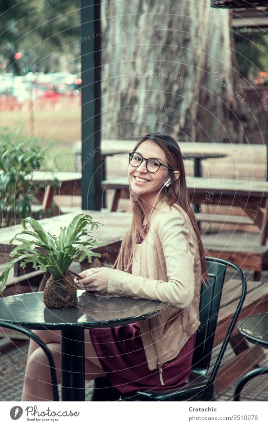 Girl with long hair and glasses in a dress sitting on a terrace using her cell phone and listening to music vertical nature tree palm garden mobil cellphone