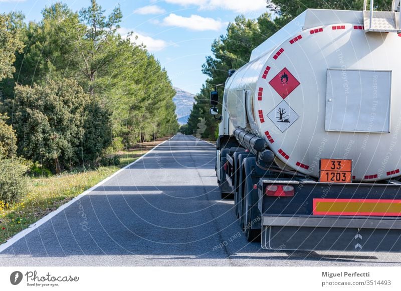 Hazard labels for flammable and polluting liquid on a fuel tank truck driving on a narrow, straight road lined with trees. road transport dangerous liquids
