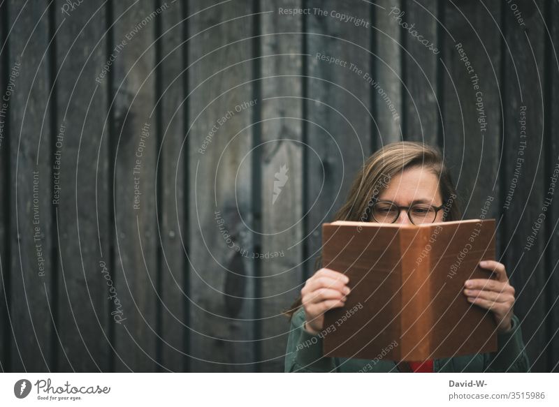 young woman with glasses reading a book outside in front of a rustic wooden wall Woman one Book Reading books Eyeglasses Wooden wall Rustic conceit formed