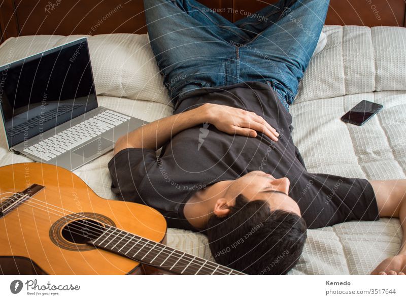 Bored man sleeping in a big bed after playing guitar and using a laptop to stay busy. Concept of spend time at home with music and technology. tire rest casual