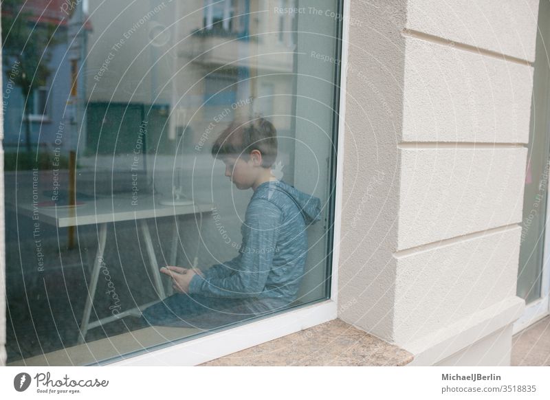 Boy sits lonely at the window and looks at his mobile phone during lockdown isolation in the coronavirus epidemic Boy (child) Child Lonely Loneliness by oneself