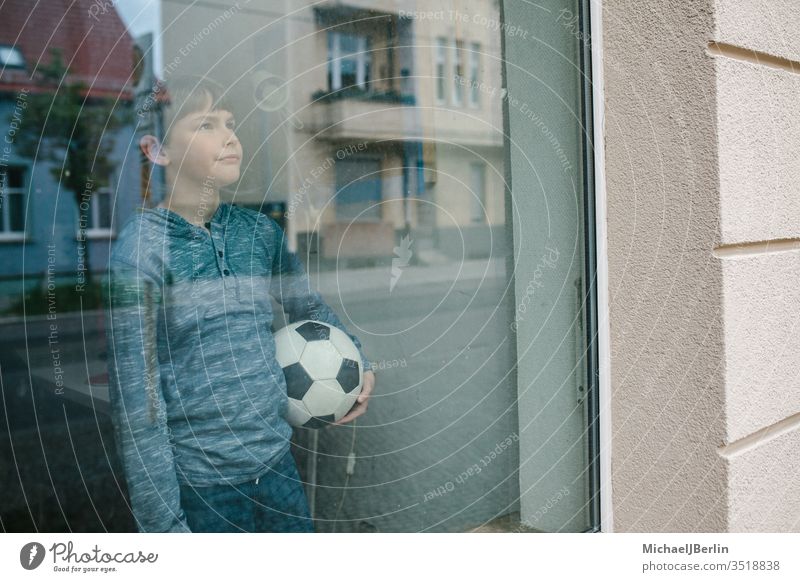 Boy stands at the window with a football and looks sadly outside, symbolizing the isolation of children during Corona Pandemic Child Boy (child) soccer inside