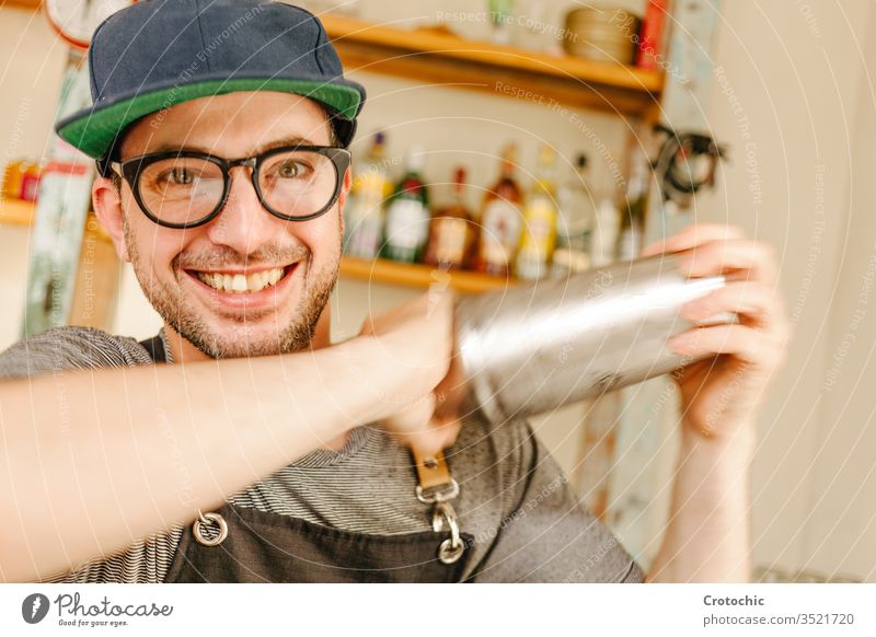 Waiter wearing an apron, glasses and a cap smiling while shaking a cocktail alcohol club fingers hold job party professional service shake bar liquor making