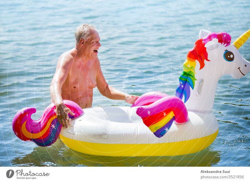 Elderly couple having fun on inflatable flamingo and unicorn. Funny active pensioners happy together enjoying summer vacation on the beach in Europe, laughing, playing the fool, splashing water.