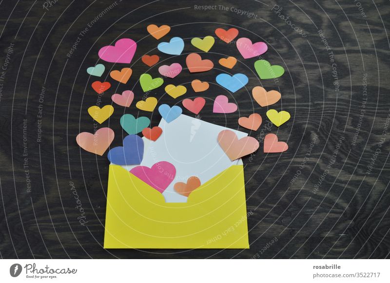 yellow envelope opened with a blank white card for self labelling, many colourful hearts cut out of paper which come out of it as a nice greeting from a distance with social distance e.g. for Mother's Day or birthday on black wood