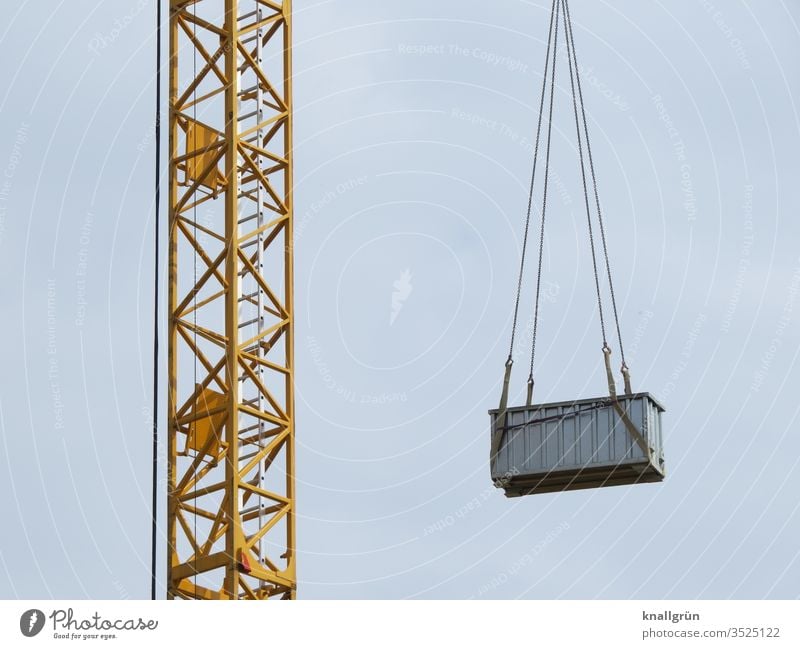 Yellow construction crane with a medium-sized grey metal container suspended from chains on the jib Crane Container hang Construction site Construction crane