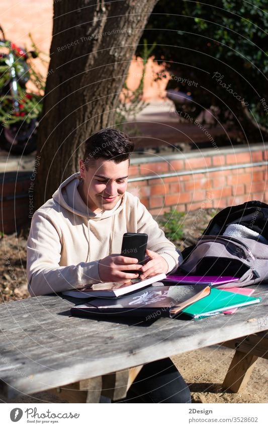 Young student sitting in a park while using a mobile phone on a wooden table one person communication technology education computer notebook studying internet
