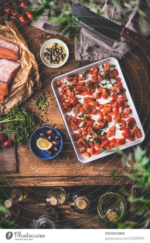 Cooking in a vintage kitchen.  Tomatoes on a baking sheet, raw fish , herbs and seasonings, olives oil , cleaver on a rustic wooden table background. Top view. Mediterranean food concept