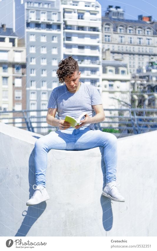 Man with a modern hairstyle sitting on a white wall reading a book vertical city urban seat park street sunny sunbath summer relax holiday stylish fashion man