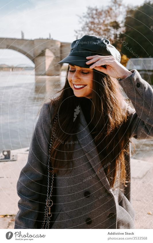 Cheerful young woman on embankment river smile style outerwear adjust cap happy female trendy quay pier eyes closed water street city shore coast urban cheerful