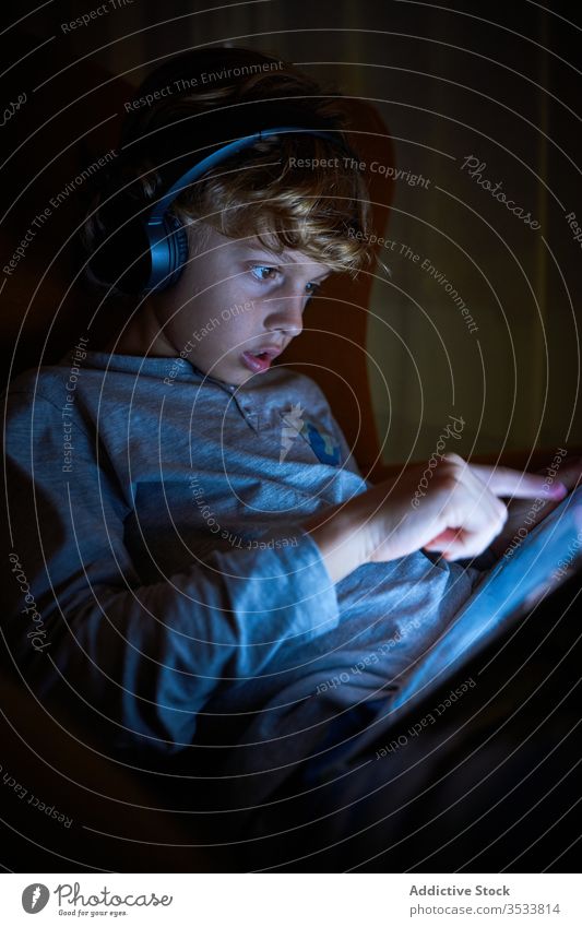 Vertical photo of a child sitting with headphones on a sofa touching the screen of a tablet at night boy pointing interact imagination cinema lateral multimedia