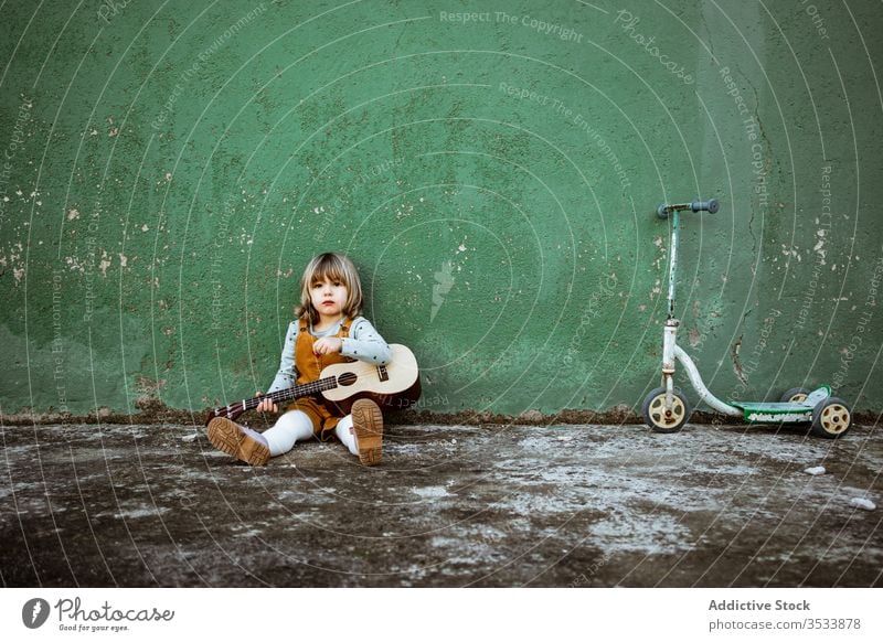 Girl with ukulele sitting near shabby wall girl music kick scooter play casual little rough ground weathered green child street kid building grunge instrument