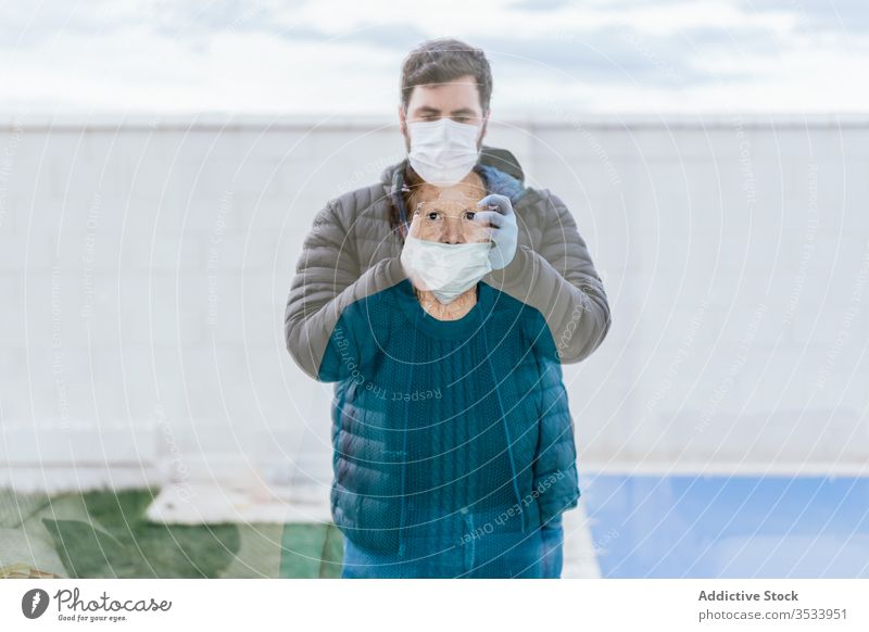 Young guy in medical mask taking picture of aged woman in medical mask through glass grandmother quarantine self isolation separate take photo social distancing