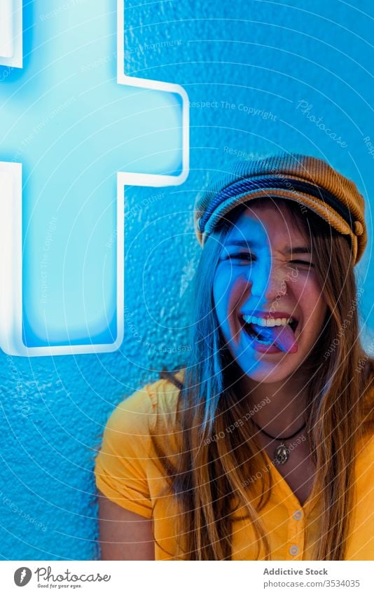 Young cheerful woman making funny face on blue background show tongue neon rebel tongue out make face having fun grimace sign yellow trendy wall medical cross